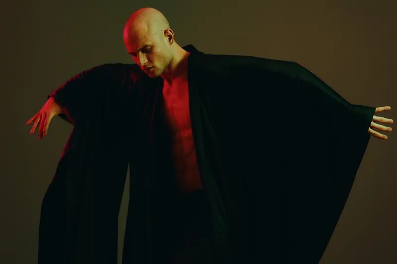 Artistic portrayal of a man in a dramatic pose, wearing a flowing black garment, showcasing a mix of shadows and vibrant lights, enhancing the emotional depth of the scene.
