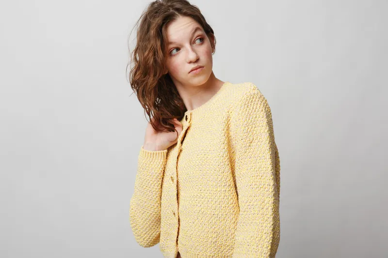 Young woman with curly hair wearing yellow knitted sweater looks sideways with thoughtful expression. Studio portrait on grey background. Concept of casual fashion and natural beauty.