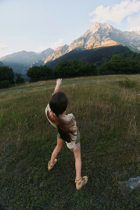 A young woman with long dark hair reaches out toward a mountain peak while standing in a grassy field during a clear day in nature.