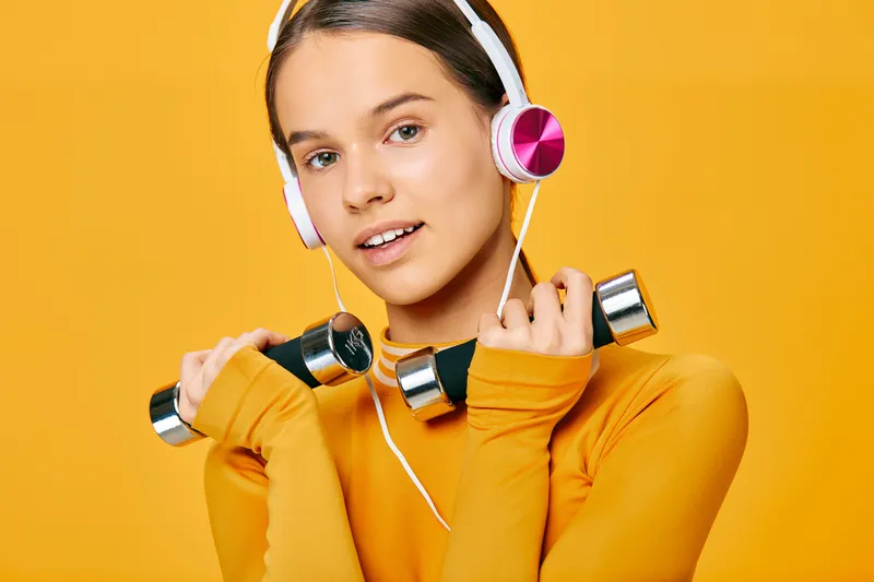 Woman in bright orange sweater wearing headphones lifts small dumbbells in a fitness moment, expressing sport and active lifestyle with focused training and colorful studio backdrop.