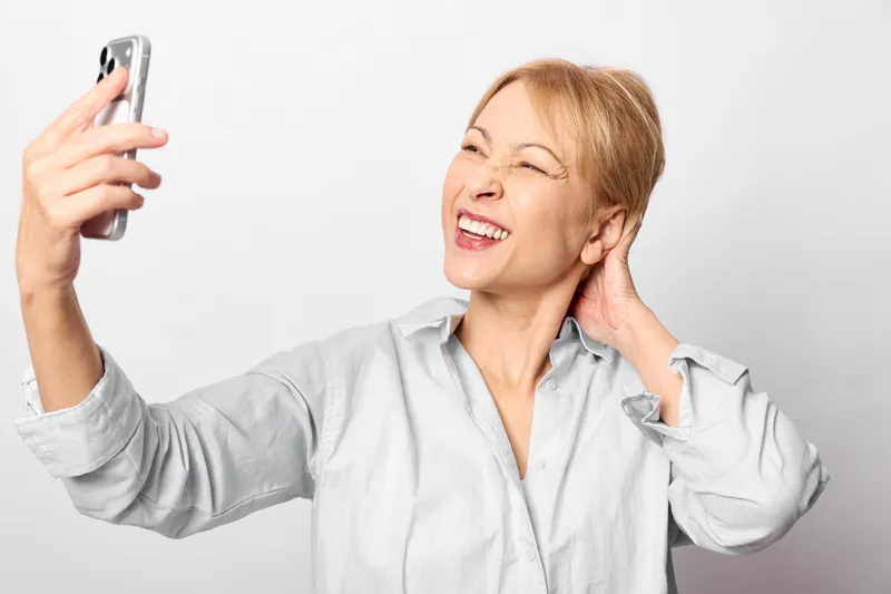 Smiling elderly woman takes a selfie with a smartphone, portrait on a clean bright background. Hand at her ear suggests communication. Concept of technology and relaxed lifestyle.