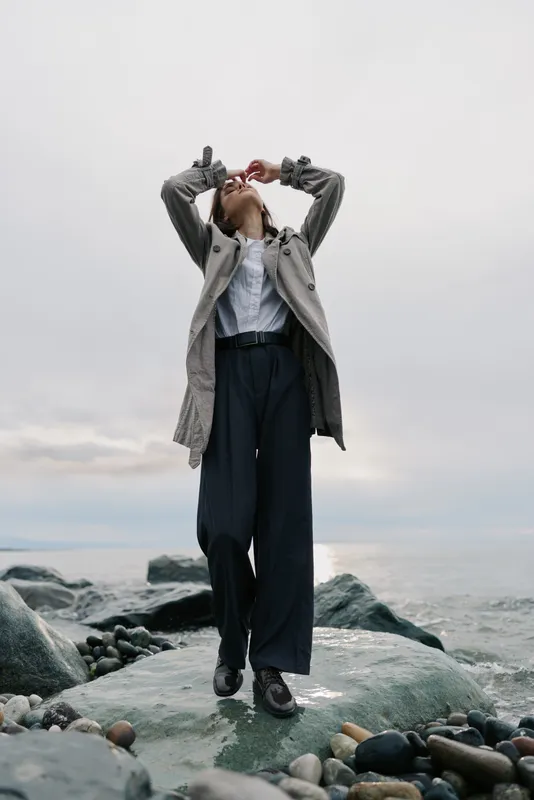Fashionable woman in grey coat and black trousers stands on rocky shore near sea under cloudy sky. Casual style, outdoor portrait, natural light, autumn mood, freedom and confidence concept.