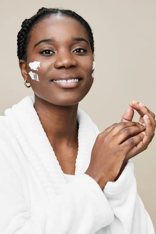 Happy Black woman applying moisturizer in a cozy bathrobe, showcasing healthy skin care routine and self care concept with a soft neutral background