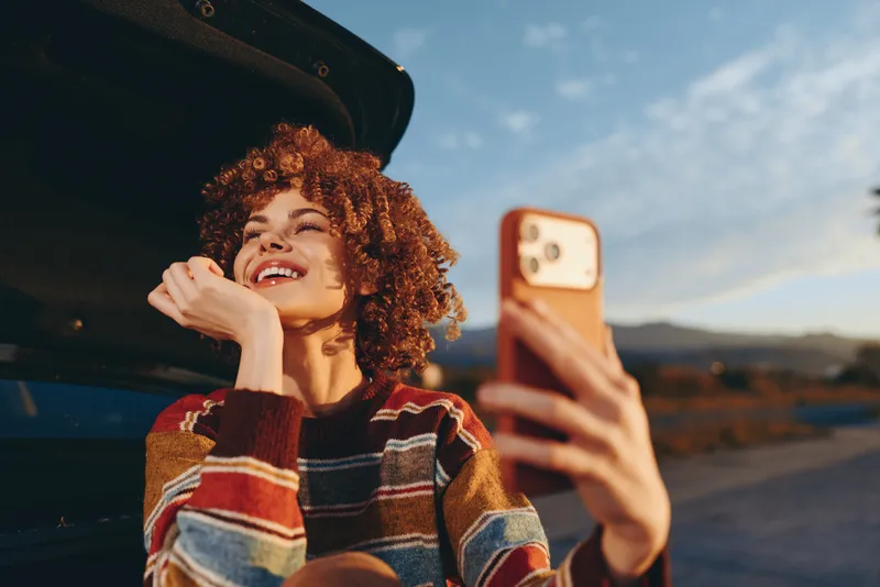 Cheerful woman lifestyle outdoors near a car, laughing with curly hair in warm sunset light, holding a phone for selfie, relaxed posture, natural colors, scenic road, evening