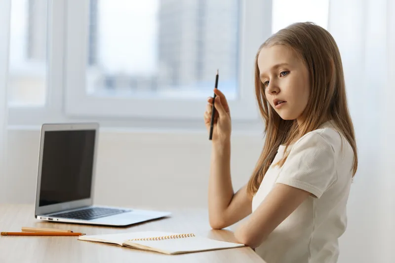Thoughtful girl with pencil in hand, seated at a desk with a laptop, reflecting on her studies in a bright, modern office environment