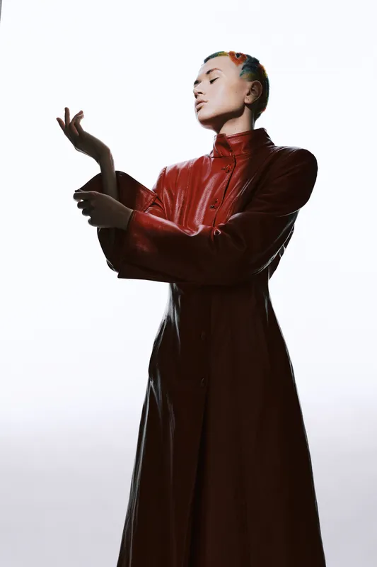 young woman in red leather coat posing gracefully with closed eyes against white background, contemporary fashion style with strong dramatic lighting