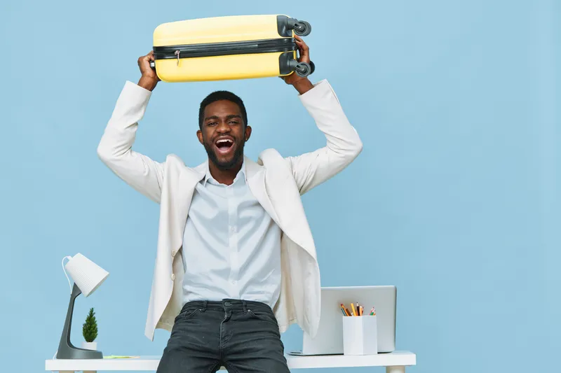 Cheerful young African American man in business casual attire holding a yellow suitcase overhead, standing in a modern office with blue background, expressive and joyful mood, concept of travel, succe