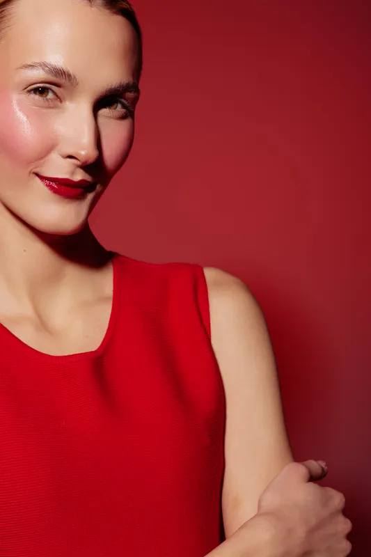 Smiling woman with shiny skin and red lipstick wears sleeveless red dress against red background. Portrait of confident female model with natural makeup and smooth complexion in studio lighting.