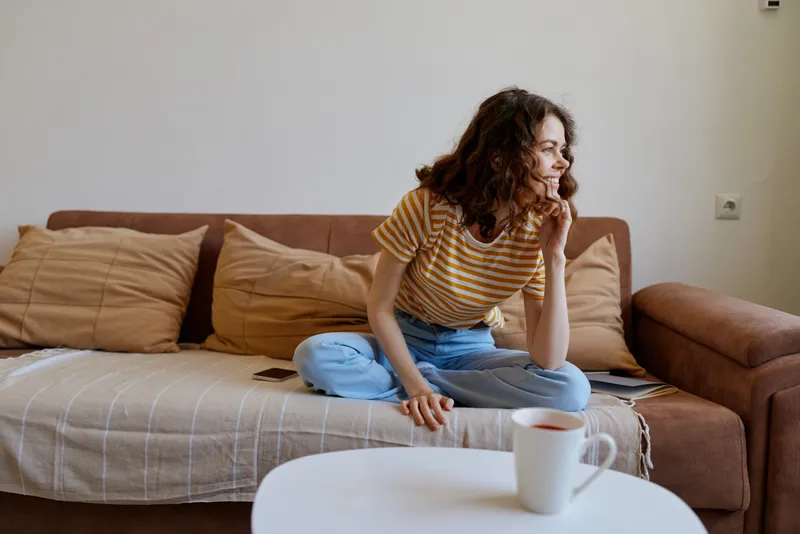 A pensive woman sits on a cozy sofa at home, leaning forward with a thoughtful gaze. A warm cup rests on a small table, capturing a moment of quiet reflection.