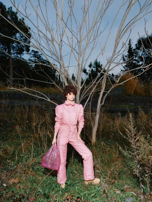Woman wearing a pink jumpsuit poses beside a bare tree in a grassy field, carrying a pink handbag, dramatic lighting, autumn mood, casual chic outdoor fashion shoot with natural backdrop.