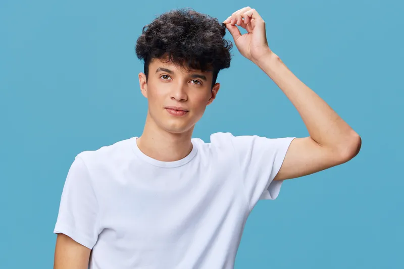 young man in white t-shirt poses confidently against light blue background, casual style, curly hair, studio portrait with soft lighting