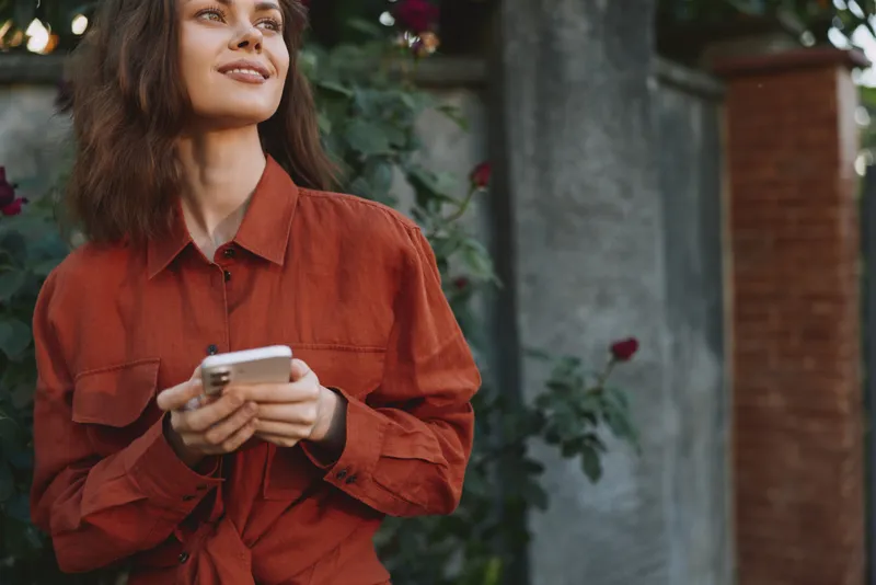 Cheerful woman outdoors, looking upward, smiling, holding smartphone, casual red shirt, sunlit garden backdrop, relaxed lifestyle, natural colors, lush greenery, candid moment
