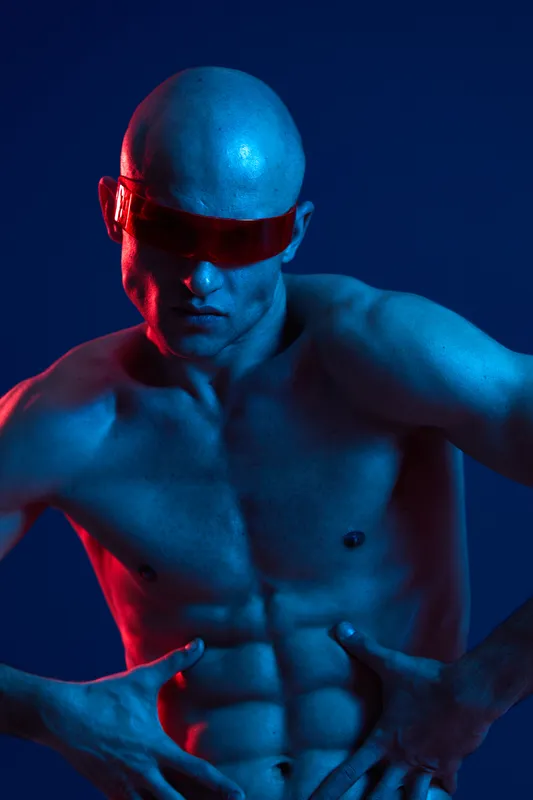 Strong man with sunglasses poses dramatically, showcasing his athletic physique against a blue and red background. The striking hues create a unique visual impact.