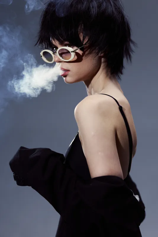 Stylish woman with short hair wearing sunglasses exhales smoke in a dark minimalistic setting with a moody atmosphere