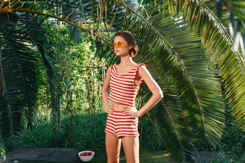 A cheerful woman in a striped red and white outfit stands in a lush garden, smiling and confident under bright sunlight, creating a playful and relaxed summer atmosphere.