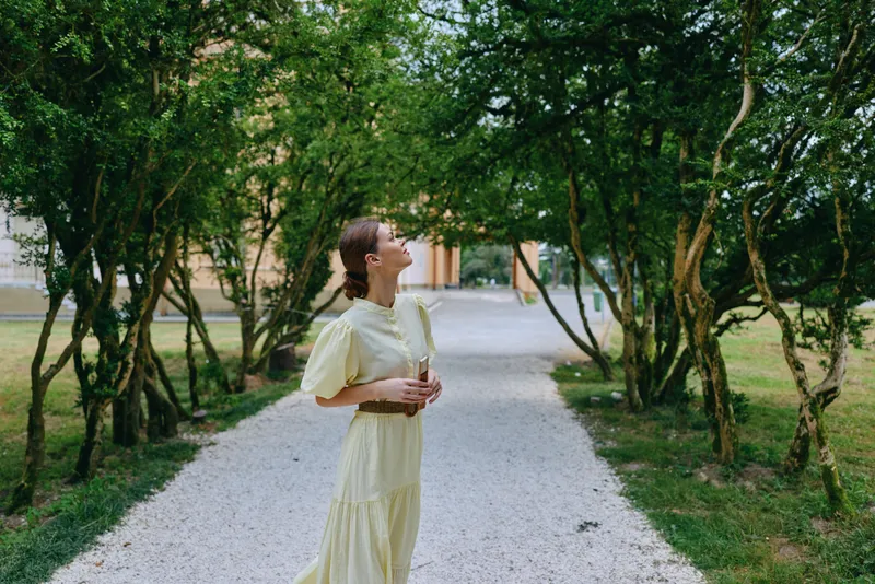 Woman in a pale yellow dress stands on a gravel path, gazing upward among leafy trees in a peaceful outdoor park setting, radiating calm and contemplation