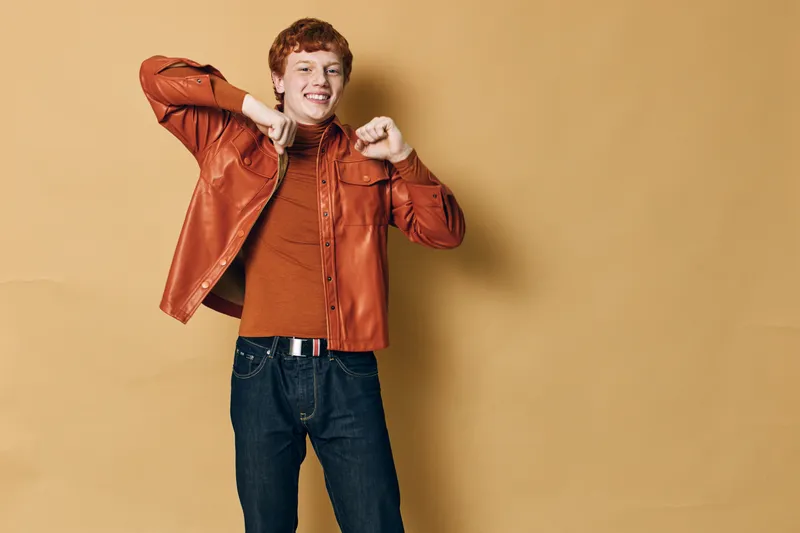 Young man with red hair wearing orange leather jacket and sweater posing smiling against beige background. Casual style, happy male portrait with denim jeans and belt buckle in studio.