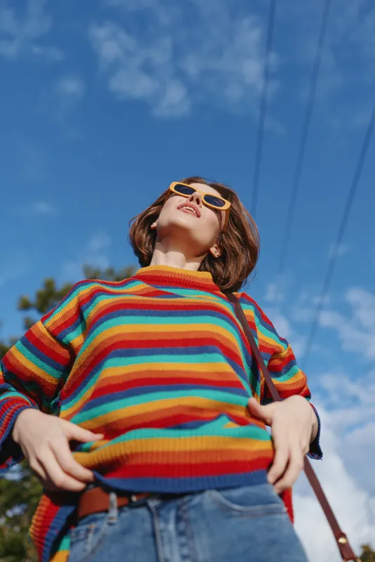 Woman in colorful striped sweater and sunglasses looks up in low angle portrait against blue sky outdoors. Casual fashion, confidence, youth and vibrant street style.