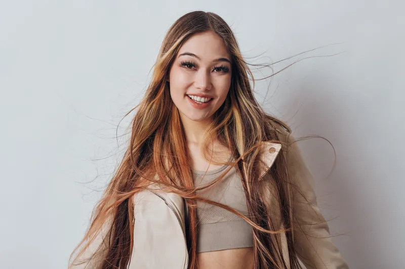 Young woman with long hair smiling and wearing casual clothes standing against plain background. Blonde girl with happy expression and wind blowing through hair portrait.