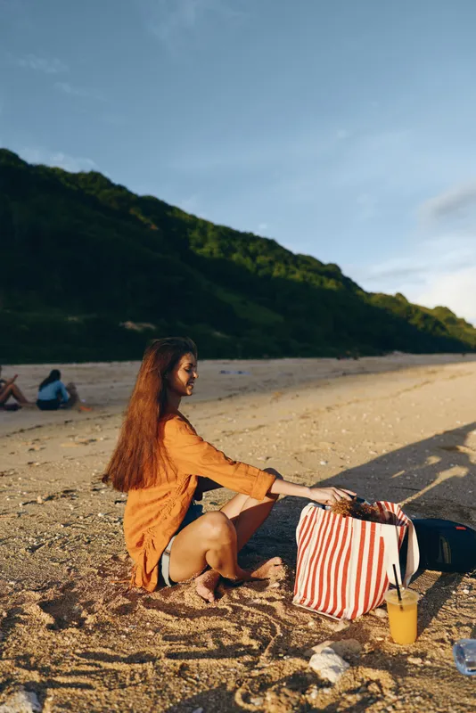 Woman on beach in summer sunny weather, relaxation and travel mood, sitting on sand and holding a striped bag, ocean horizon behind, outdoor nature landscape, calm trip concept today