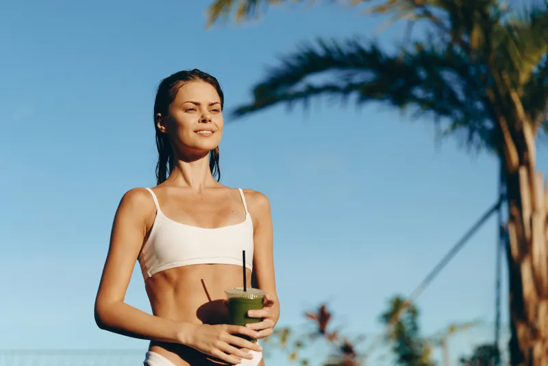 Bright outdoor scene with a smiling woman in a light bikini top, holding a green smoothie under warm sun, palm trees in the background, relaxed summer lifestyle.