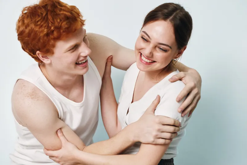 Two young people smiling and hugging each other in casual white tank tops on light background. Friends sharing joyful moment with laughter and close embrace together.