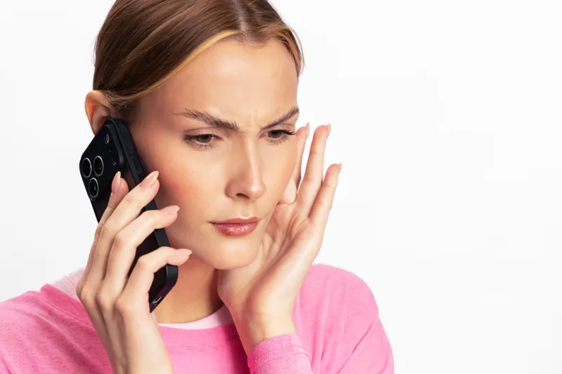 Stressed young woman in pink sweater holds smartphone to ear and touches face with concerned expression. Isolated on white background. Concept of anxiety, communication, and worry.