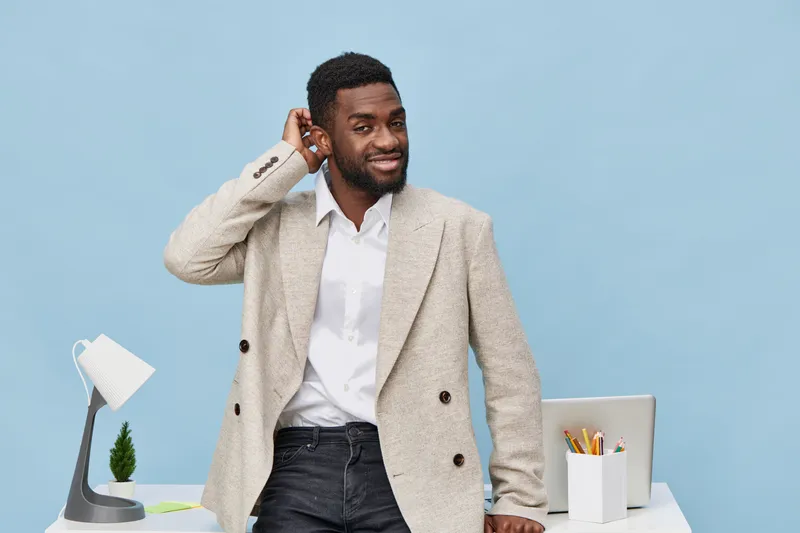 Confident young African American man in beige blazer adjusting his hair while standing in modern office setup with white desk, laptop, and desk lamp against light blue background. Business casual styl