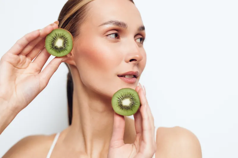 Bright portrait of a woman holding kiwi slices near her face, skincare inspired moment, soft lighting, natural glow, clean background, fresh beauty concept, playful