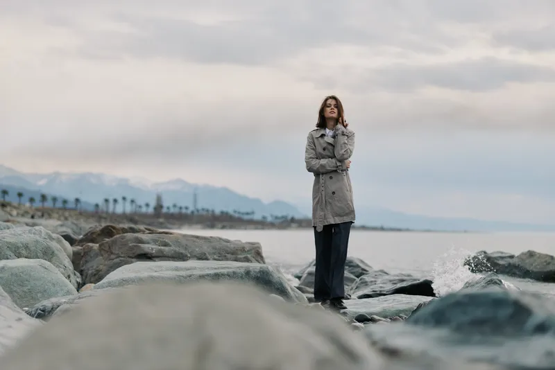 A confident woman stands on rocky shore wearing a grey coat and dark pants. The cloudy sky and distant mountains create a calm, contemplative coastal scene.