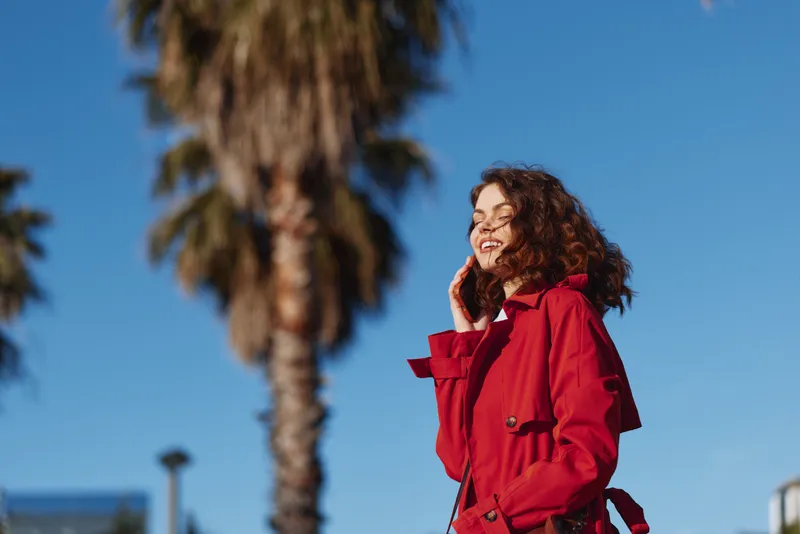 A smiling woman in a red coat stands outdoors under a clear blue sky, palm trees in the background. A bright fashion moment focused on confident style.