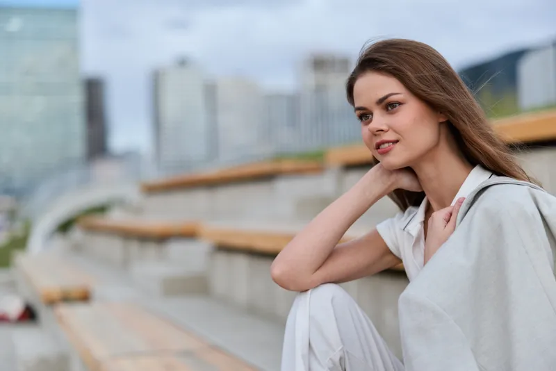 Young woman in white casual wear sitting thoughtfully outdoors with cityscape background, natural light and calm expression in spring fashion style