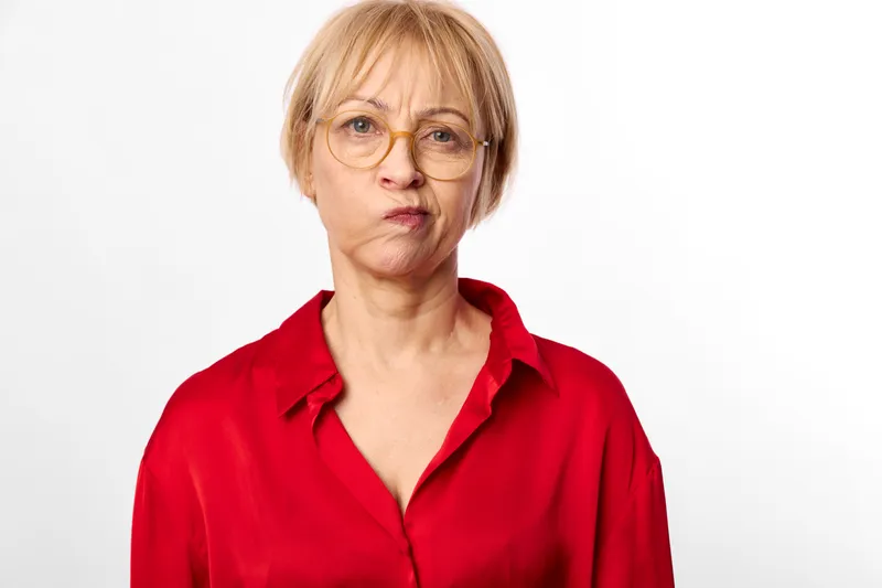 Portrait of an older woman with glasses, serious concerned expression in a red blouse, plain white background, she conveys worry and stress during aging, studio headshot concept