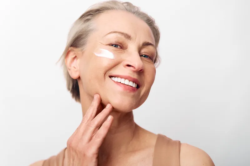Smiling middle aged woman applies white face cream on cheek, showing healthy glowing skin and natural beauty in soft studio light. Concept of skincare, self care, and wellness routine.