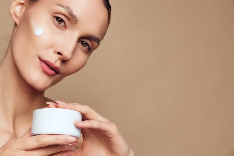 Young woman with clear skin holding white jar of cream applying moisturizer on cheek, natural makeup, soft lighting, neutral background, skincare beauty routine concept