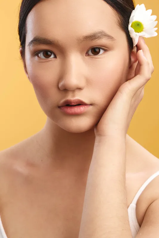 asian woman posed in a graceful close up portrait, flower tucked behind her ear, soft studio lighting, calm expression, and clean background.