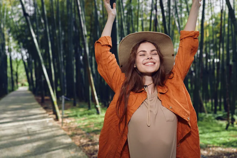 Woman smile hat bamboo forest path, joyful young female with raised arms on sunlit trail, wearing orange jacket and straw hat, portrait of freedom, travel and outdoor relaxation, walking on paved