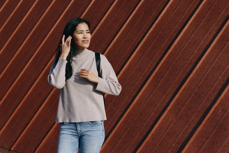 Casual woman with backpack walking beside a diagonal wooden wall in a knit sweater and jeans, capturing a street style moment for lifestyle photography.