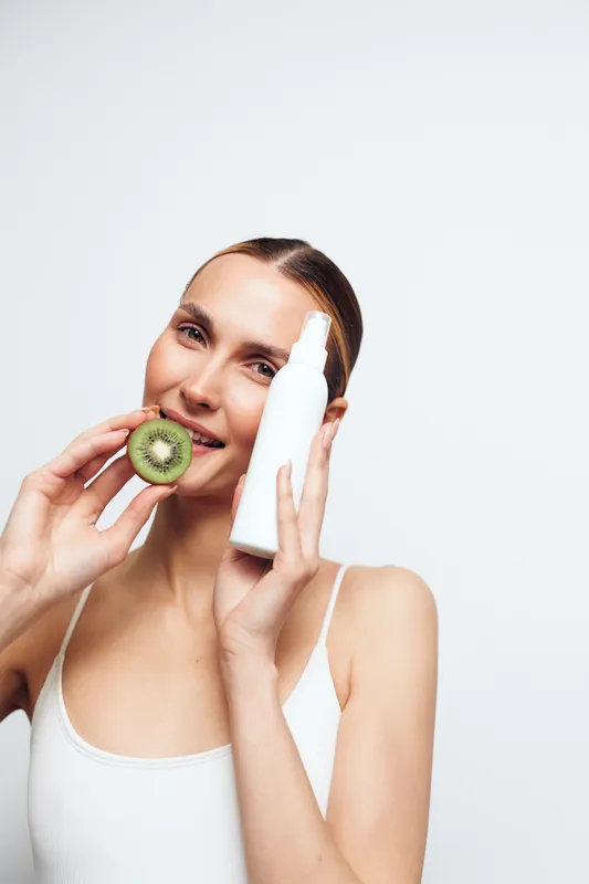 Young woman with slicked back hair holds a white cosmetic bottle and a sliced kiwi while smiling against a white background in a skincare concept.