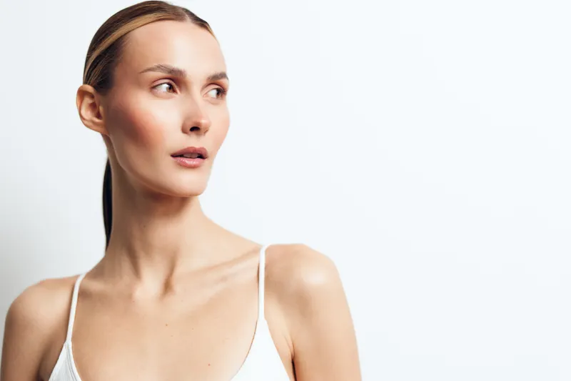 A thoughtful young woman in a white tank top gazes off to the side in a bright studio, conveying introspection and quiet confidence.