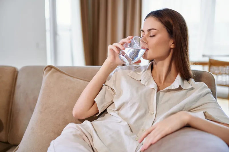 Woman drinking water from a glass on a sofa at home, relaxed and hydrated in casual loungewear, portrait lifestyle scene showing comfort, wellness and everyday self care.