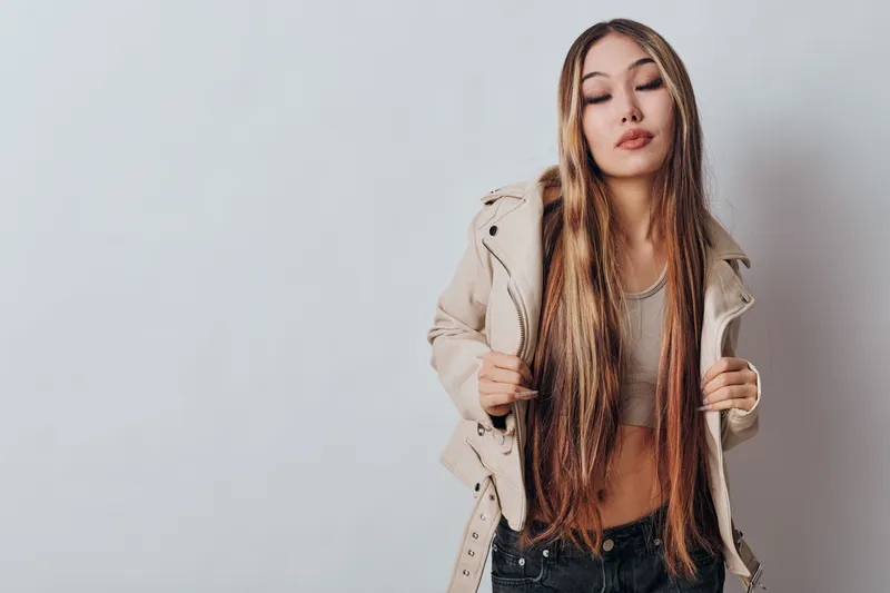 Young woman with long hair and eyes closed posing in fashionable beige jacket and casual outfit on plain background for style and beauty lifestyle portrait.