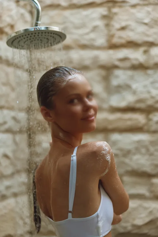 woman showering outdoors under rain shower with wet hair and relaxed expression, natural light highlighting smooth skin and white swimwear strap