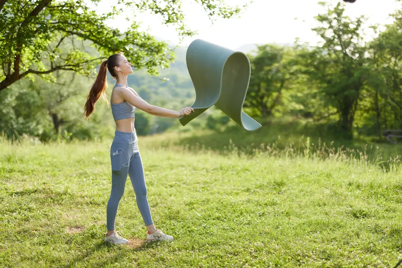 Young woman practicing yoga outdoors in athletic wear, engaged in a mindful moment with her mat in a sunny green field, embodying wellness and serenity