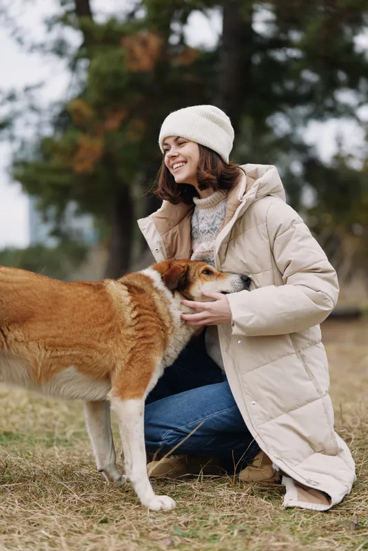Woman outdoors in a warm winter coat kneels on grass, gently embracing a dog in a park, showing affection and companionship during a chilly day for a candid lifestyle shot.