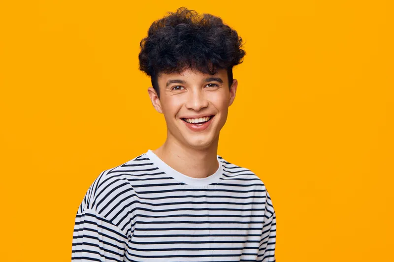 smiling young man wearing a striped shirt stands against an orange background, showing happy expression and casual style in bright studio lighting.