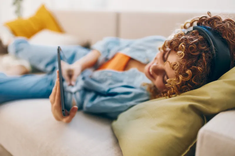 Счастливая женщина держит планшет и наушники, наслаждаясь музыкой на уютном диване дома
