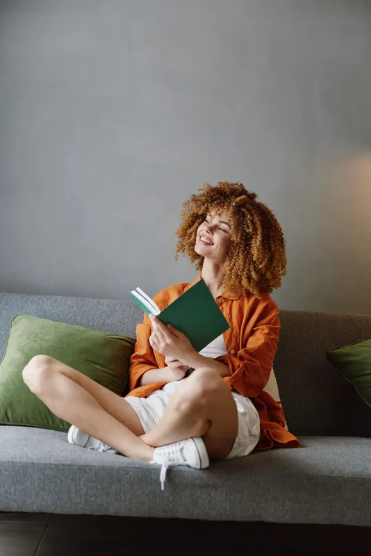 Молодая женщина с вьющимися волосами с удовольствием читает книгу на зелёном диване, одетая в повседневную одежду и выражающая радость