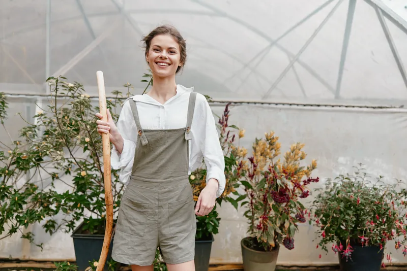 Young woman wearing overalls in a greenhouse, smiling joyfully while holding a gardening tool, surrounded by lush green plants and vibrant flowers