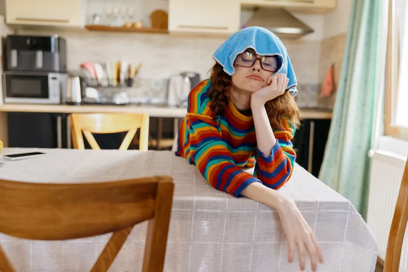 Young woman with curly hair rests her chin on her hand at a kitchen table, wearing a colorful striped sweater and a blue beanie, looking thoughtful.